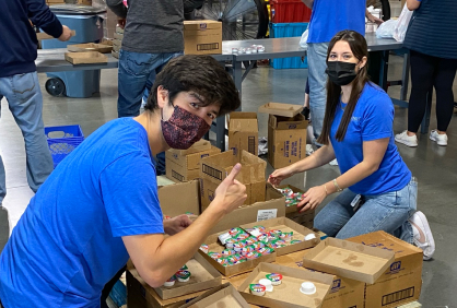 Two Kainos employees wearing protective masks and blue volunteer shirts crouched over boxes of donation supplies